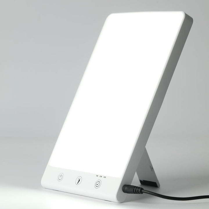 The Fashiongirl Light Therapy Lamp 10,000 lux LED is a rectangular lamp with a bright white screen, fold-out support, three control buttons, and a power cord at the bottom—designed to simulate artificial daylight.