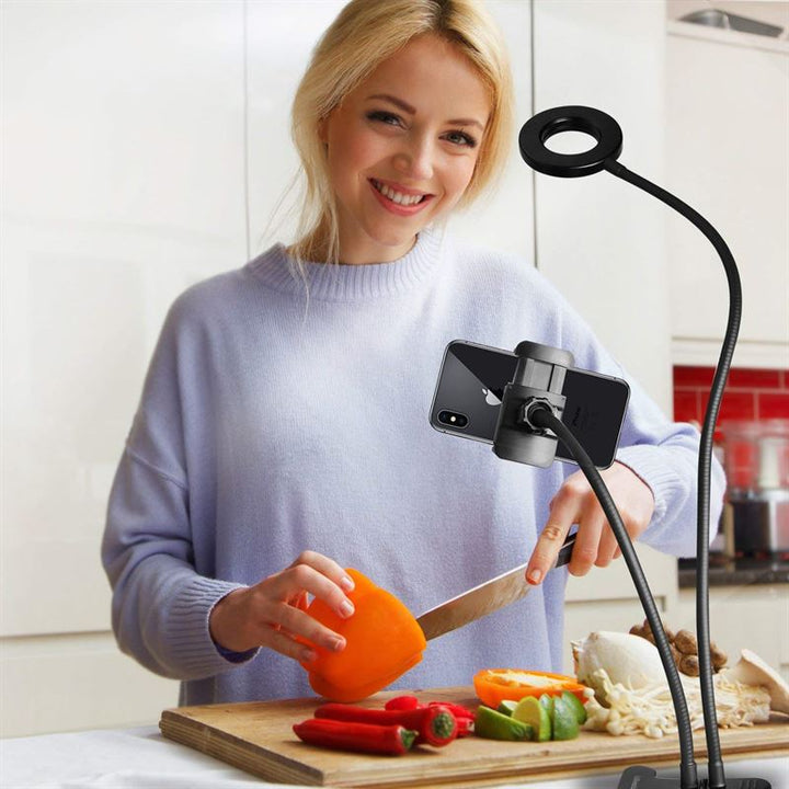 A woman slices an orange bell pepper in her kitchen, filming with the UNIQ Selfie Ring Light with LED brightness control and flexible arms, ensuring steady, well-lit shots on her smartphone for streaming or vlogs.