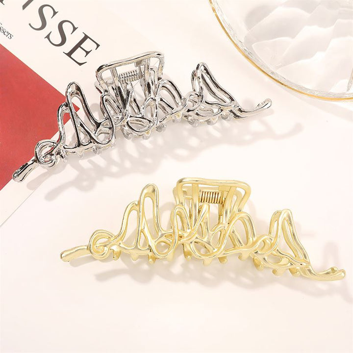Die SOHO Asli Metal Hair Claw in Silber von SOHO zeichnet sich durch ein wellenförmiges, abstraktes Design in Metallic-Optik aus und wird auf einer hellen Oberfläche neben einer klaren Glasschale und einer Zeitschrift präsentiert – perfekt für jede stilvolle Haaraccessoire-Kollektion.