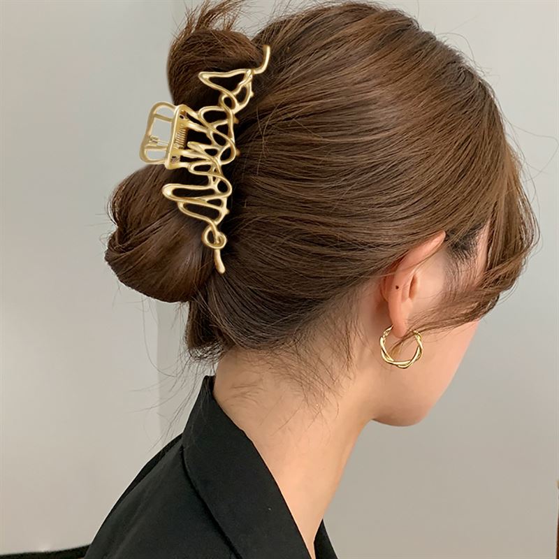 Eine Frau mit braunen Haaren, die zu einem tief sitzenden Dutt gesteckt sind, der mit der SOHO Asli Metal Hair Claw – Silver von SOHO befestigt ist. Sie trägt ein schwarzes Oberteil und goldene, gedrehte Creolen. Das Foto, das von hinten und von der Seite aufgenommen wurde, hebt ihr elegantes silbernes Haaraccessoire hervor.