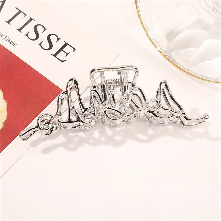 Die SOHO Asli Metal Hair Claw in Silber von SOHO zeichnet sich durch ein modernes, wellenförmiges Design aus und glänzt stilvoll neben einer Glasschale und einer Zeitschrift mit dem Schriftzug „MATISSE” – ein schickes und zeitgemäßes Accessoire.