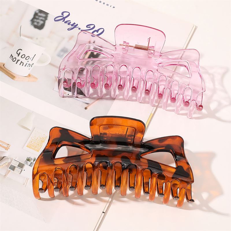 Two large SOHO Astrid Hair Claws, one in brown tortoise and one in translucent pink, are displayed on an open magazine. A white mug with "Good morning" printed on it is seen in the background.