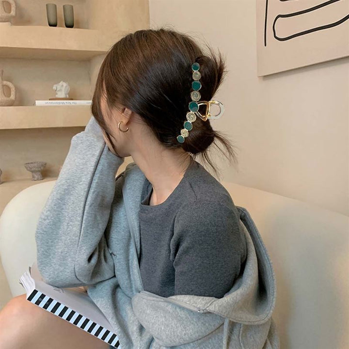 A woman with dark hair in a secure messy bun, held by the SOHO Becca Hair Claw - Midnight, sits on a couch in a grey sweater and holds a striped notebook. Her face is turned away from the camera.
