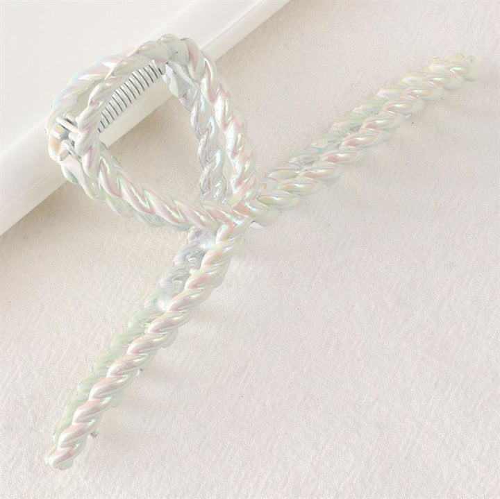 The SOHO Caprina Hair Claw - White by SOHO features an iridescent glazed chrome finish and a twisted rope bow design, shown resting on a light-colored textured surface.