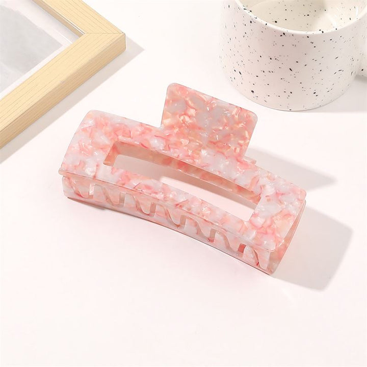 The SOHO Clara Hair Claw in Light Coral (XL) rests on a white surface beside a speckled white cup and a wooden picture frame.