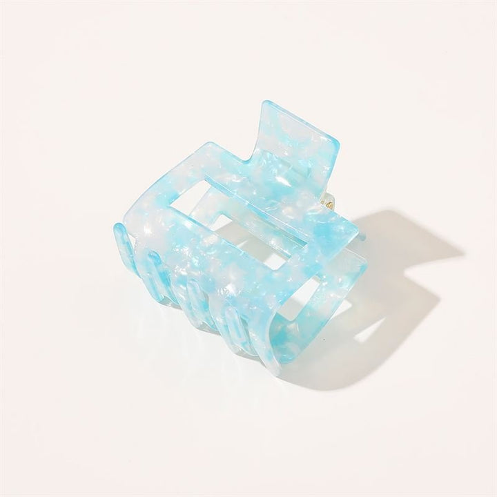 Die SOHO Ema Hair Claw in Crystal Blue von SOHO zeichnet sich durch ein durchscheinendes blaues Marmormuster aus, das auf weißem Hintergrund mit einem weichen Schatten dargestellt wird – ein schickes Accessoire, ideal für elegante Frisuren.