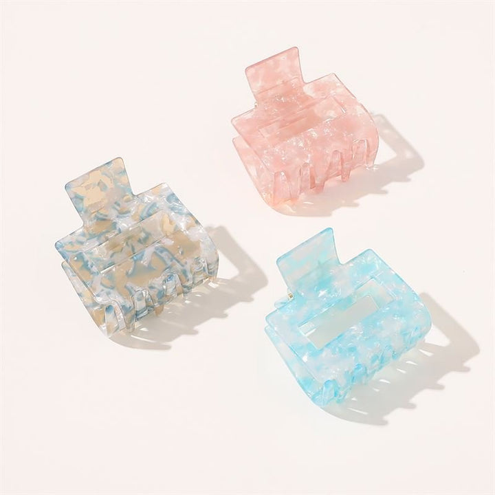 Die SOHO Ema Hair Claw – Crystal Blue von SOHO hat ein durchscheinendes, marmoriertes, rechteckiges Design mit breiten Zinken und eignet sich perfekt für elegante Frisuren. Die Clips werden neben Pastellrosa und -grün auf einem hellen Hintergrund präsentiert.
