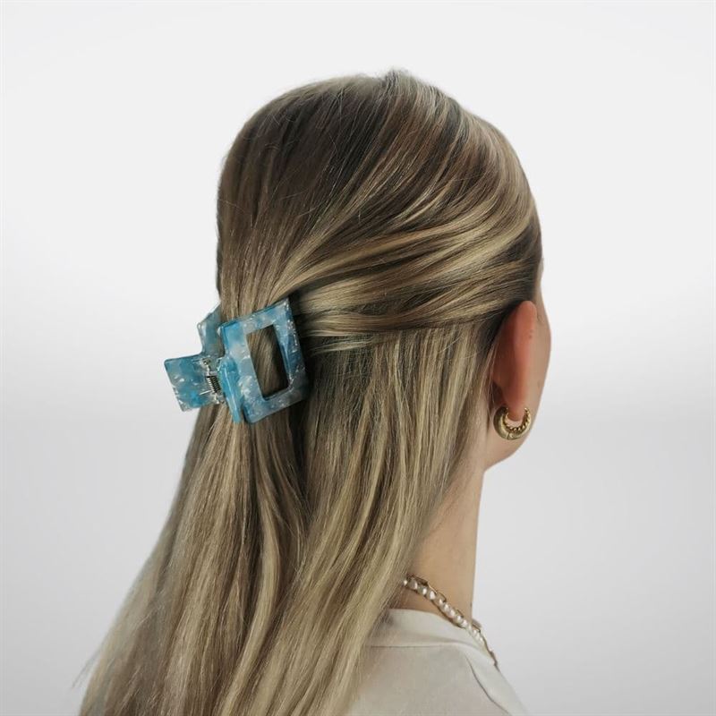 Eine Person mit langen, blonden Haaren ist von hinten zu sehen, wobei der obere Teil mit der SOHO Ema Hair Claw in Crystal Blue befestigt ist. Goldene Creolen und eine Perlenkette runden den stilvollen Look ab und präsentieren dieses schicke SOHO-Accessoire.
