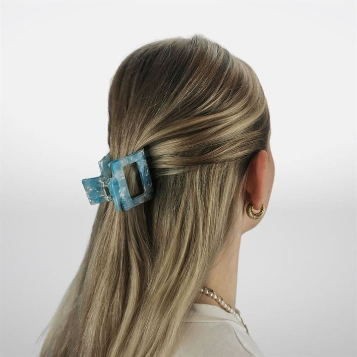 Eine Person mit langen, blonden Haaren ist von hinten zu sehen, wobei der obere Teil mit der SOHO Ema Hair Claw in Crystal Blue befestigt ist. Goldene Creolen und eine Perlenkette runden den stilvollen Look ab und präsentieren dieses schicke SOHO-Accessoire.