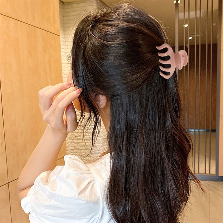 Eine Frau mit langen dunklen Haaren, von hinten gesehen, verwendet die SOHO Esta Hair Claw – Green, um ihr Haar teilweise hochzustecken und zusätzlichen Halt zu erhalten. Sie trägt ein weißes Oberteil und steht in einem Innenraum.