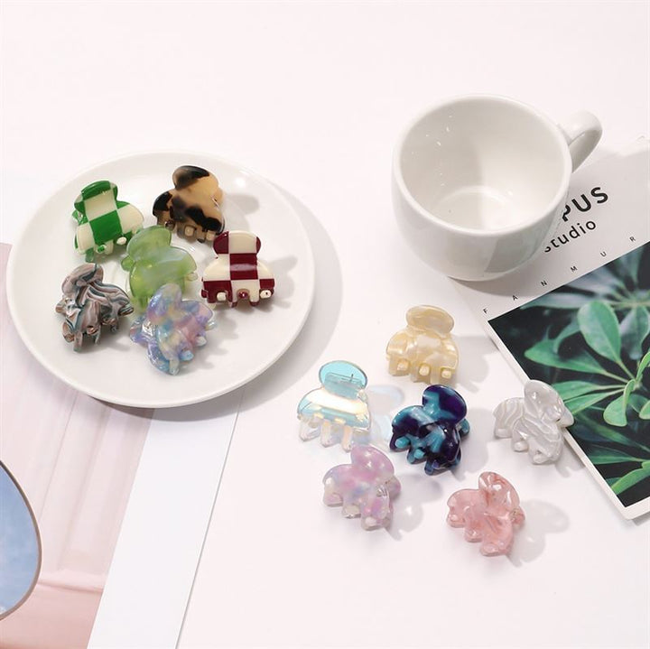 The SOHO Hara Mini Hair Claw - Blush by SOHO, in various playful shapes, is displayed on a white plate next to a cup and magazine; these mini claws provide a secure hold, with extra clips scattered nearby for convenience.