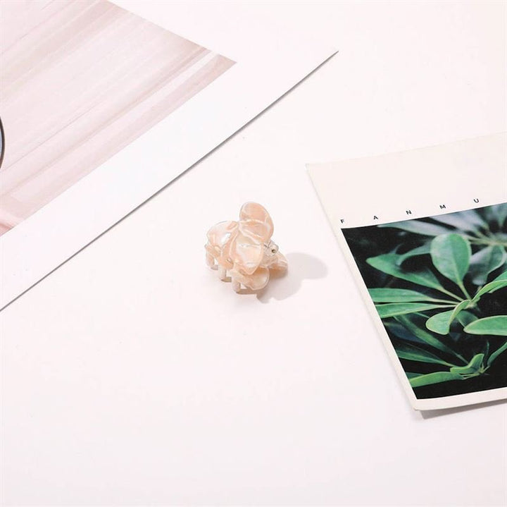 A small, light pink elephant figurine sits on a white surface beside the SOHO Hara Mini Hair Claw - Cloud Pink by SOHO, between two papers—one with a leafy green plant photo and the other with abstract light pink lines.