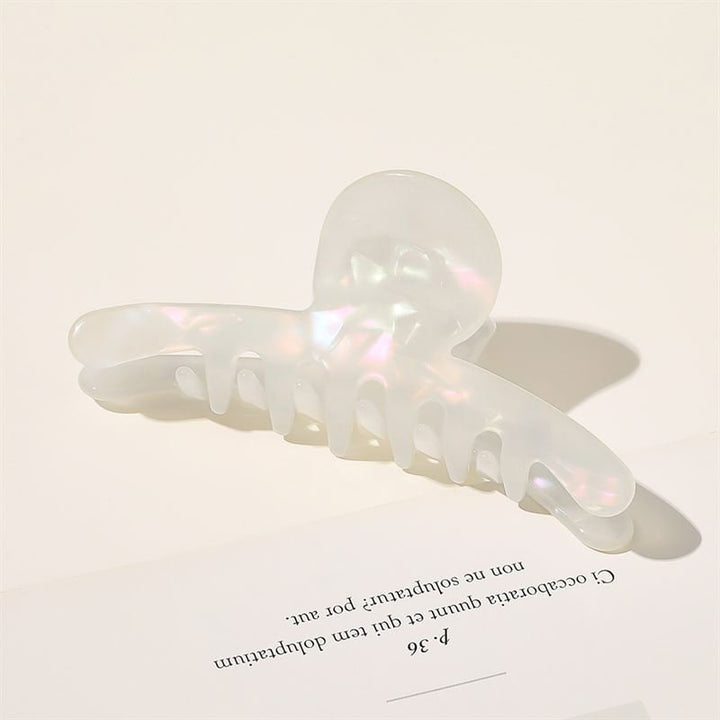 The SOHO Haylen Hair Claw in White by SOHO rests on a white surface beside a sheet of paper with partially visible printed text, casting a soft shadow.