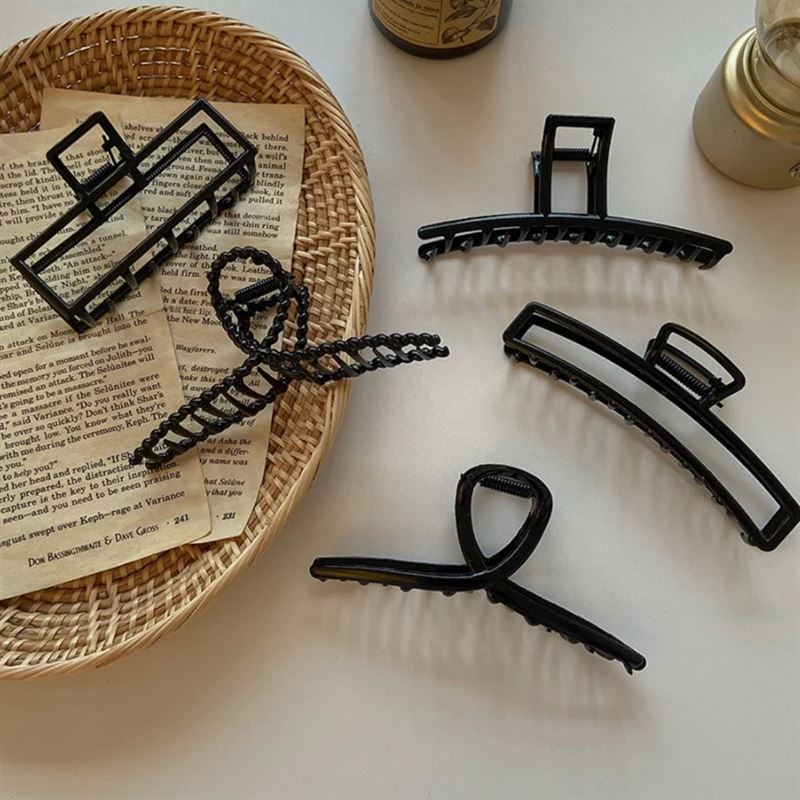 Five SOHO Iva Metal Hair Claws in black by SOHO are displayed on a white surface with a wicker tray, torn book pages, gold candle, and brown bottle—perfect for securing a messy bun or adding style like metal hair clips.