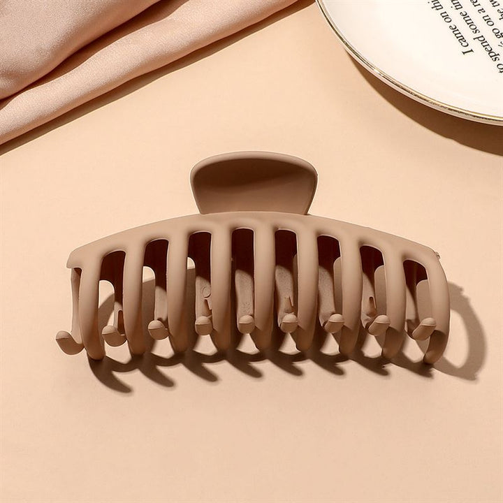 The SOHO Kana Hair Claw in beige features wide, curved prongs for a strong grip on all hair types, and is shown on a light tan surface near a white plate and beige fabric.