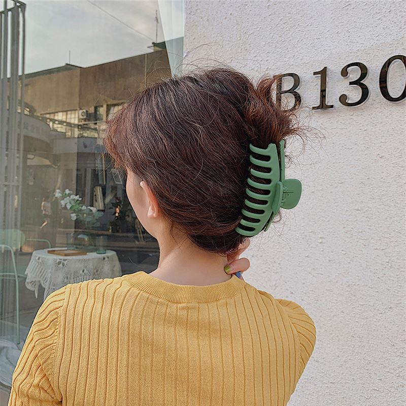 A person with wavy brown hair clipped up with the SOHO Kana Hair Claw - Beige, known for its strong grip and suitable for all hair types, stands outside facing a white wall labeled B130, wearing a mustard yellow ribbed shirt.