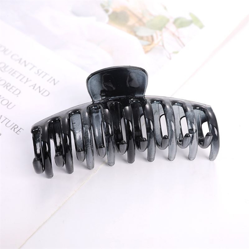 The SOHO Kana Hair Claw - Monotone Gradient by SOHO, a black plastic clip with a strong grip and curved prongs, rests on a white surface beside an open magazine showing blurred text and images.