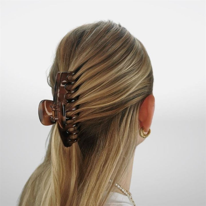 Eine Frau mit blondem Haar, das halb hochgesteckt und halb offen ist und mit der SOHO Kana Hair Claw – Tortoise befestigt ist, die für ihren starken Halt bekannt ist. Sie trägt goldene Creolen und eine Halskette, die von hinten zu sehen sind – geeignet für alle Haartypen.