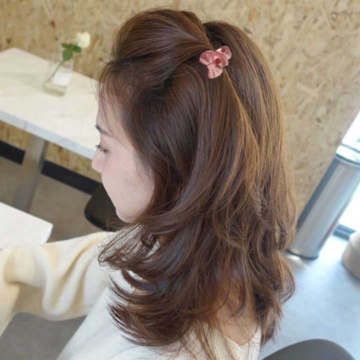 A woman with medium-length brown hair sits at a table wearing a cream sweater, her hair styled with a SOHO Lala Mini Hair Claws - Flowers by SOHO. The background features a white table, chair, and small flower vase.