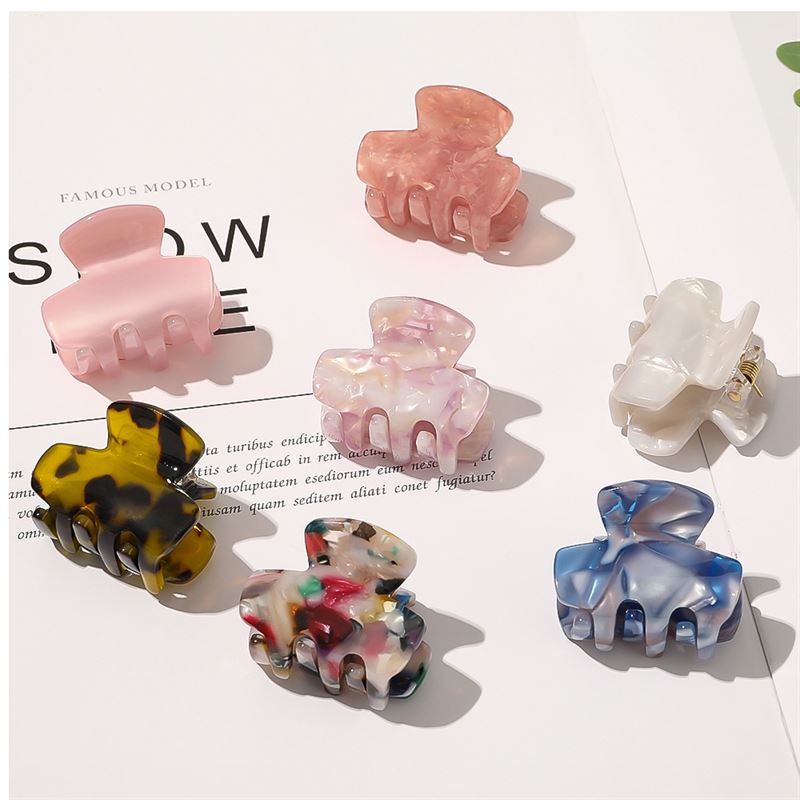The SOHO Loa Mini Hair Claw - Blush by SOHO features a pink marble-like design and comes in a set of seven colorful small clips in pink, cream, yellow, multicolor, blue, and white tones. They're arranged neatly on a white surface with a magazine beneath.