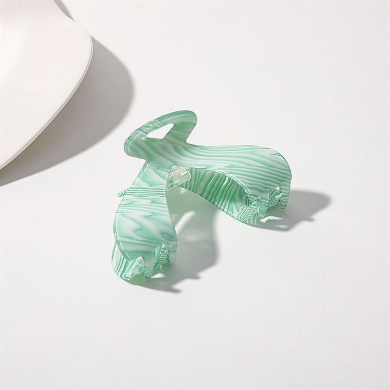 Die SOHO Malou Hair Claw – Green von SOHO liegt offen auf einer weißen Oberfläche, ihr marmoriertes grün-weißes Design wirft einen weichen Schatten. In der oberen linken Ecke erscheint ein weißes Objekt.