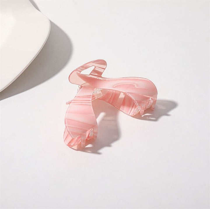Die SOHO Malou Hair Claw – Pink von SOHO mit einem durchscheinenden rosa Marmor-Design liegt auf einer weißen Oberfläche nahe dem Rand einer weißen Schale und wirft einen sanften Schatten.