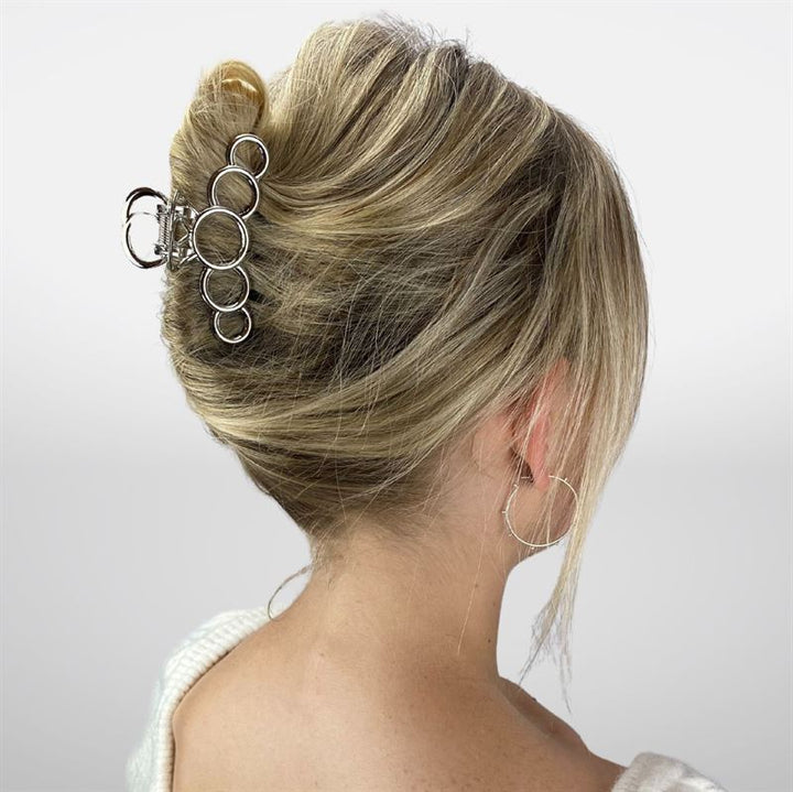 A woman with blonde hair in a French twist, secured by the SOHO Metal Hair Claw in Silver—ideal for thick hair. She wears a cream sweater and silver hoops, shown from behind against a white background.