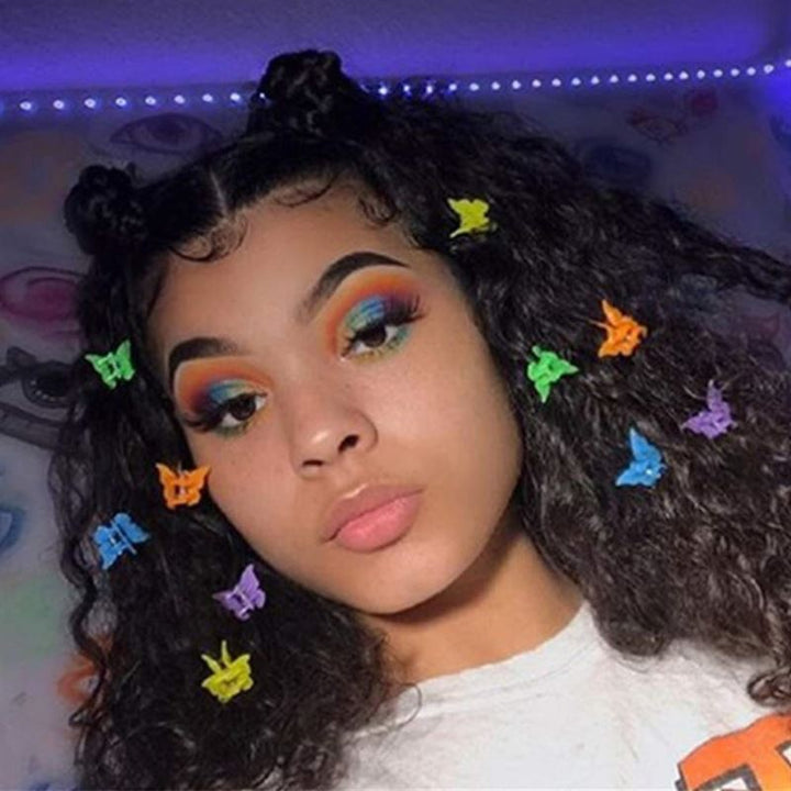 A young person with curly hair styled in two buns decorated with SOHO Mini Butterfly Hair Claws (50 pcs, multicolour) poses indoors. They wear bold blue, orange, and yellow eye makeup and a white top against an artistic wall backdrop.