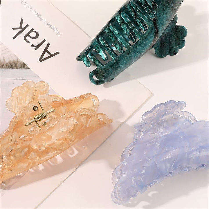 Die SOHO Naomi Hair Claw – Sunshine von SOHO verfügt über drei dekorative Klammern in durchscheinendem Pfirsich, Blau und Grün, die jedem Look eine stilvolle Note verleihen. Präsentiert auf einer weißen Oberfläche mit einem Magazin, auf dessen Cover teilweise „Arak” zu sehen ist.