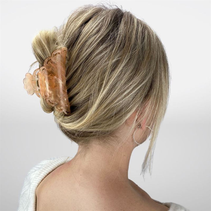 Eine Frau mit blondem Haar, das zu einem niedrigen Zopf geflochten und mit der SOHO Naomi Hair Claw – Sunshine befestigt ist, wird von hinten gezeigt. Sie trägt ein weißes Oberteil und silberne Creolen.