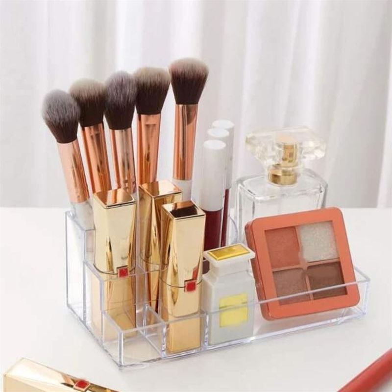 The UNIQ Acrylic Make up organiser 8 compartments by UNIQ neatly holds makeup brushes, gold lipstick tubes, lip gloss, a square eyeshadow palette, a small perfume bottle, and a compact on a white surface.
