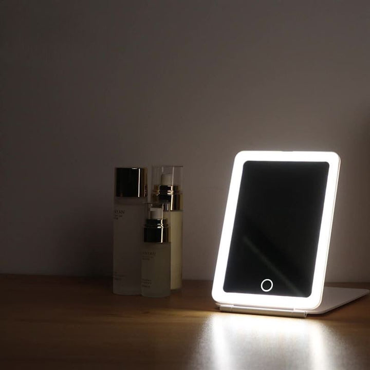 The UNIQ Foldable LED Makeup Mirror—rechargeable, travel-friendly, and white—sits illuminated on wood next to three skincare bottles against a plain wall.