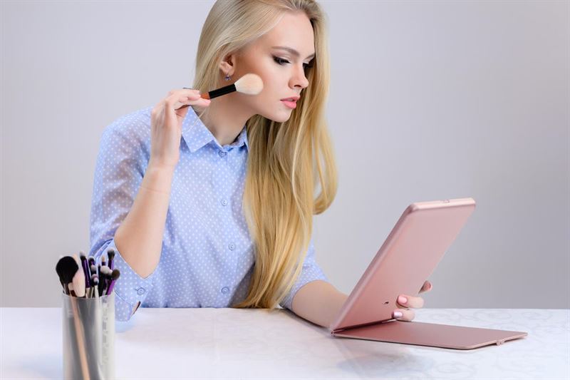 A woman with long blonde hair applies makeup using a brush while looking into the UNIQ Foldable LED Makeup Mirror by UNIQ. The rechargeable, travel-friendly white mirror sits on a white table next to a cup of brushes. She wears a blue polka dot shirt.