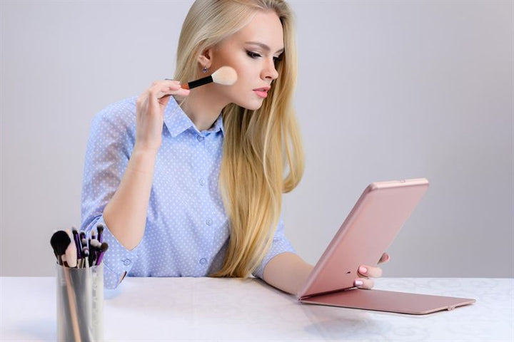 A woman with long blonde hair applies makeup using a brush while looking into the UNIQ Foldable LED Makeup Mirror by UNIQ. The rechargeable, travel-friendly white mirror sits on a white table next to a cup of brushes. She wears a blue polka dot shirt.